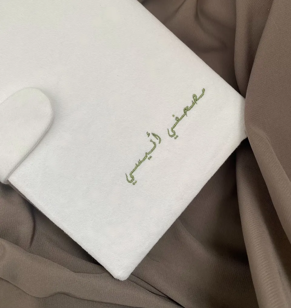White cotton book cover with rounded magnetic flap, “Mushafi anisi” embroidered in olive green Arabic calligraphy