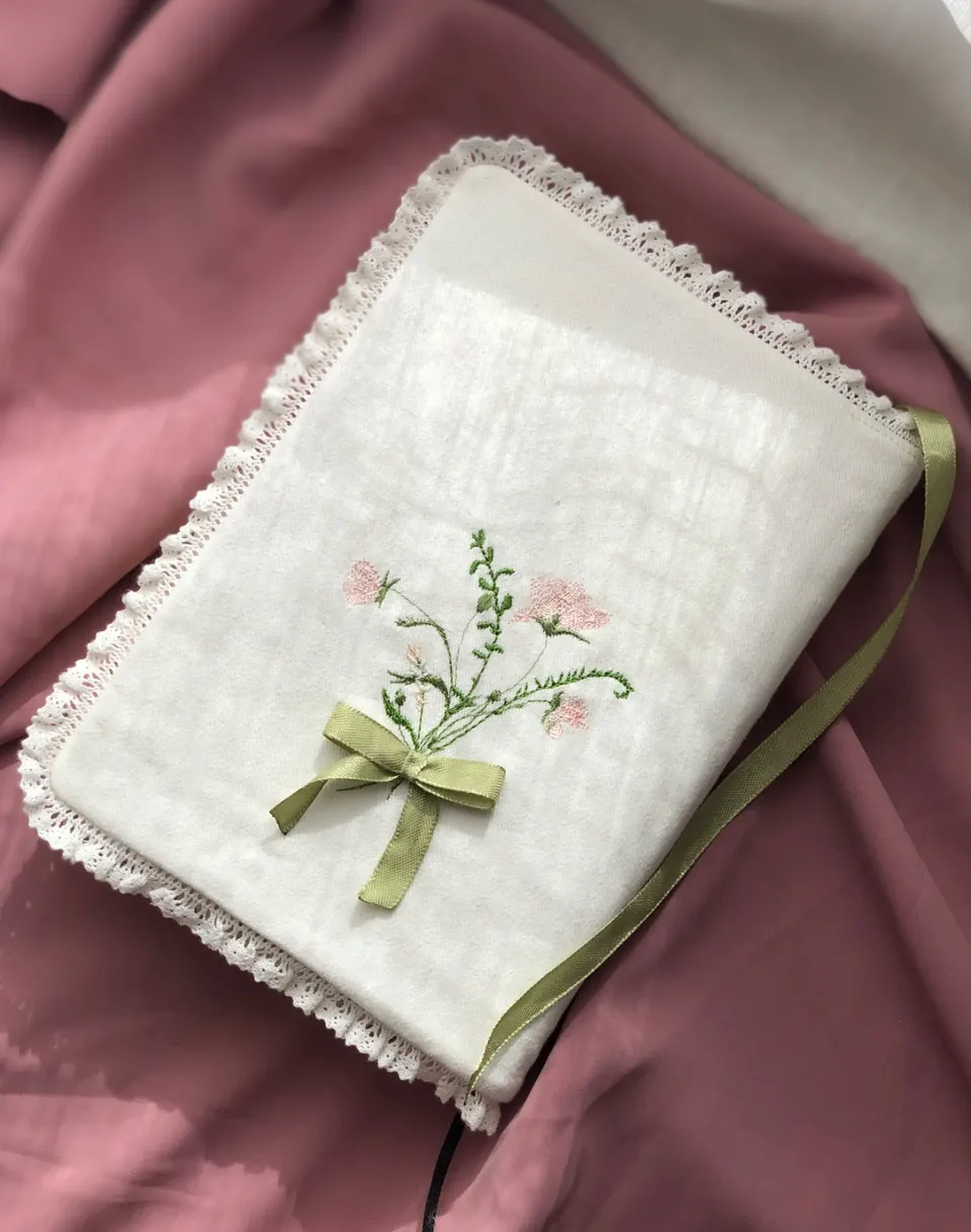 White cotton book cover, embroidered pink and green flower bouquet tied with a ribbon, white macramé lace, green ribbon bookmark