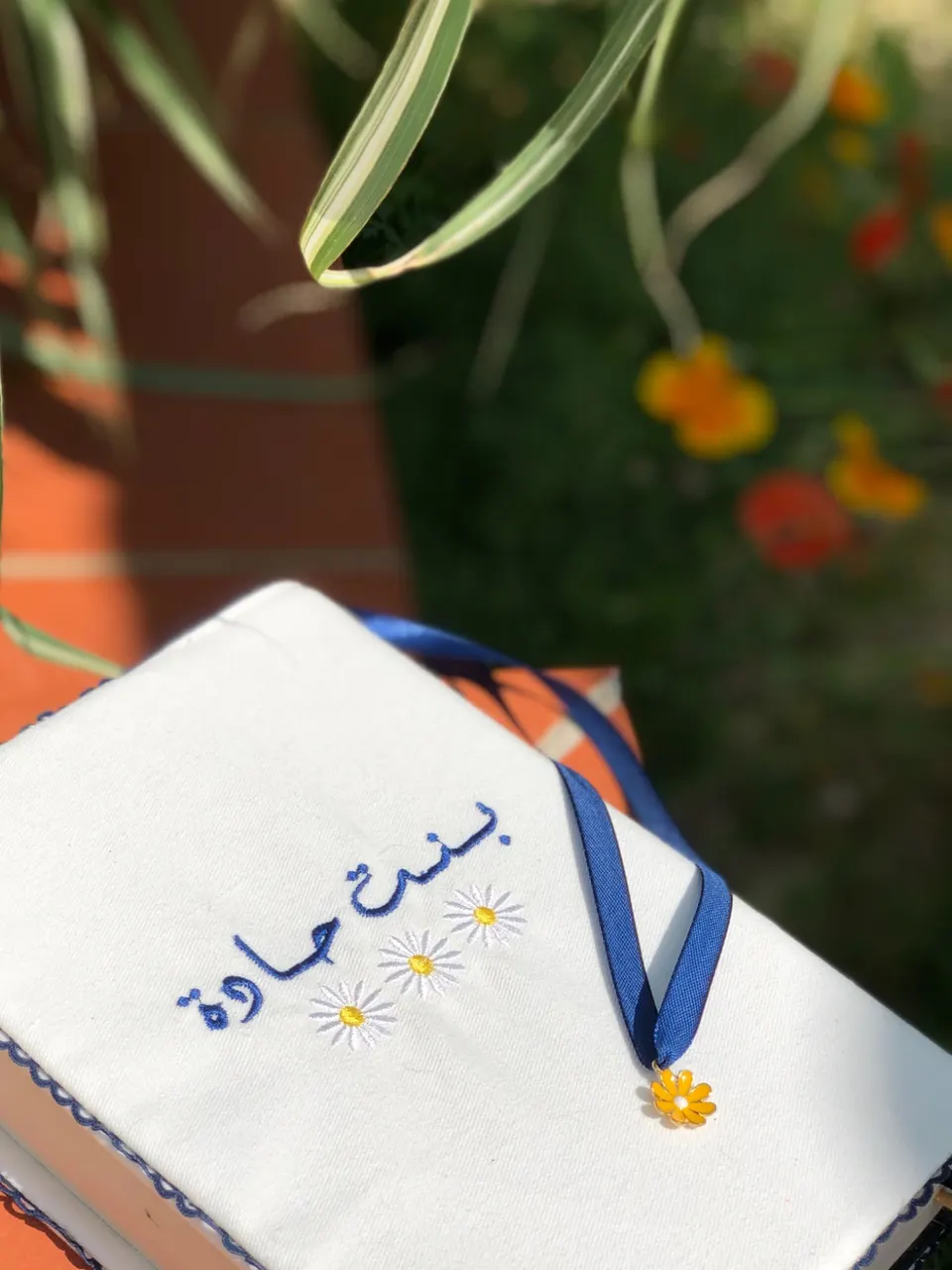 White cotton book cover, Arabic text embroidered in blue with daisies, blue ribbon with a yellow daisy charm, blue scalloped edge